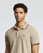 A person is wearing a Psycho Bunny MENS MONROE PIQUE POLO - B6K964D200, crafted from beige Pima cotton with a small embroidered logo on the chest. Made in Peru, the shirt features dark trim on the collar and sleeves, and its regular fit is accentuated against a plain and light-colored background.