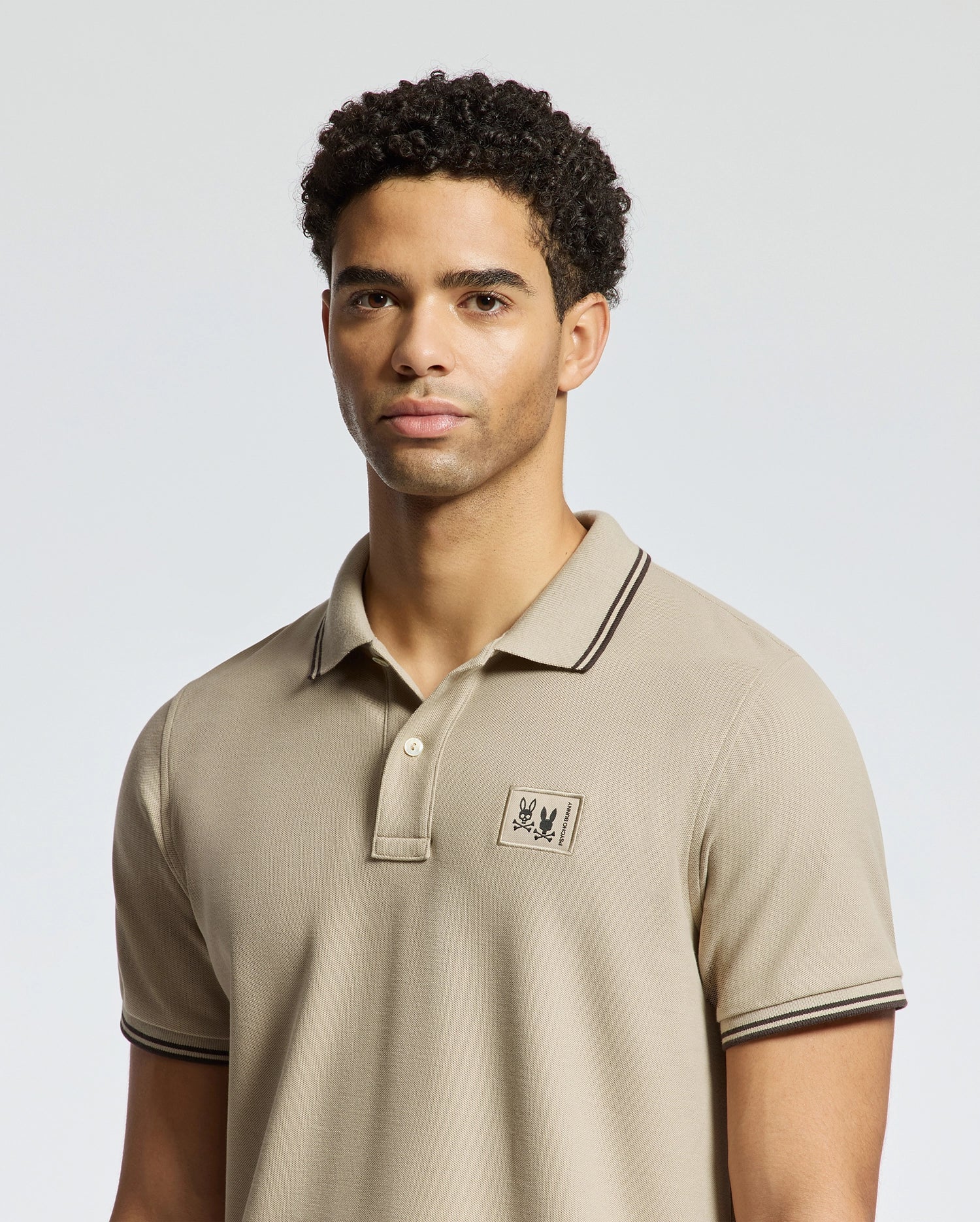 A person is wearing a Psycho Bunny MENS MONROE PIQUE POLO - B6K964D200, crafted from beige Pima cotton with a small embroidered logo on the chest. Made in Peru, the shirt features dark trim on the collar and sleeves, and its regular fit is accentuated against a plain and light-colored background.