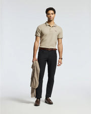A man stands against a plain background, wearing a Psycho Bunny MENS MONROE PIQUE POLO in light beige, black pants, and brown shoes. Made in Peru, the regular fit polo exudes casual elegance. He holds a light brown jacket in his left hand, his expression neutral, and his short hair styled simply.