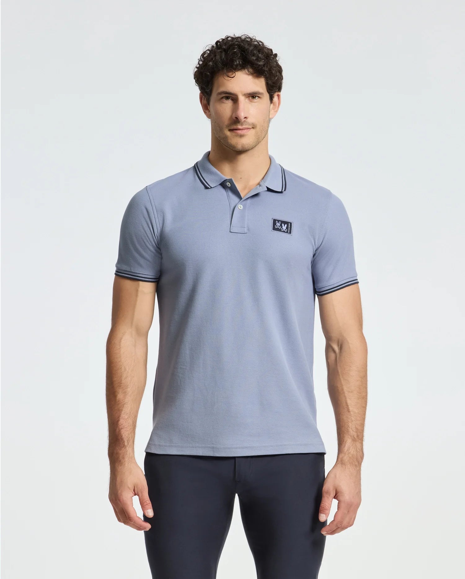 A man wearing a light blue MENS MONROE PIQUE POLO by Psycho Bunny, featuring a small emblem on the left chest, stands against a plain background. He has dark curly hair and looks directly at the camera, highlighting the regular fit of his shirt.