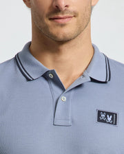 A man models the Psycho Bunny MENS MONROE PIQUE POLO - B6K964D200, made from soft light blue Pima cotton. It features an embroidered skull and crossbones on the chest, Mother-of-Pearl buttons, and a dark-trimmed collar, showcasing his upper torso and part of his face.