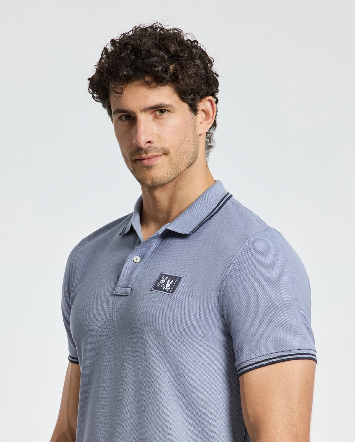 A curly-haired person is wearing a Psycho Bunny light blue MENS MONROE PIQUE POLO - B6K964D200, featuring a small logo on the chest and elegant Mother-of-Pearl buttons in a regular fit. The background is plain and light-colored.