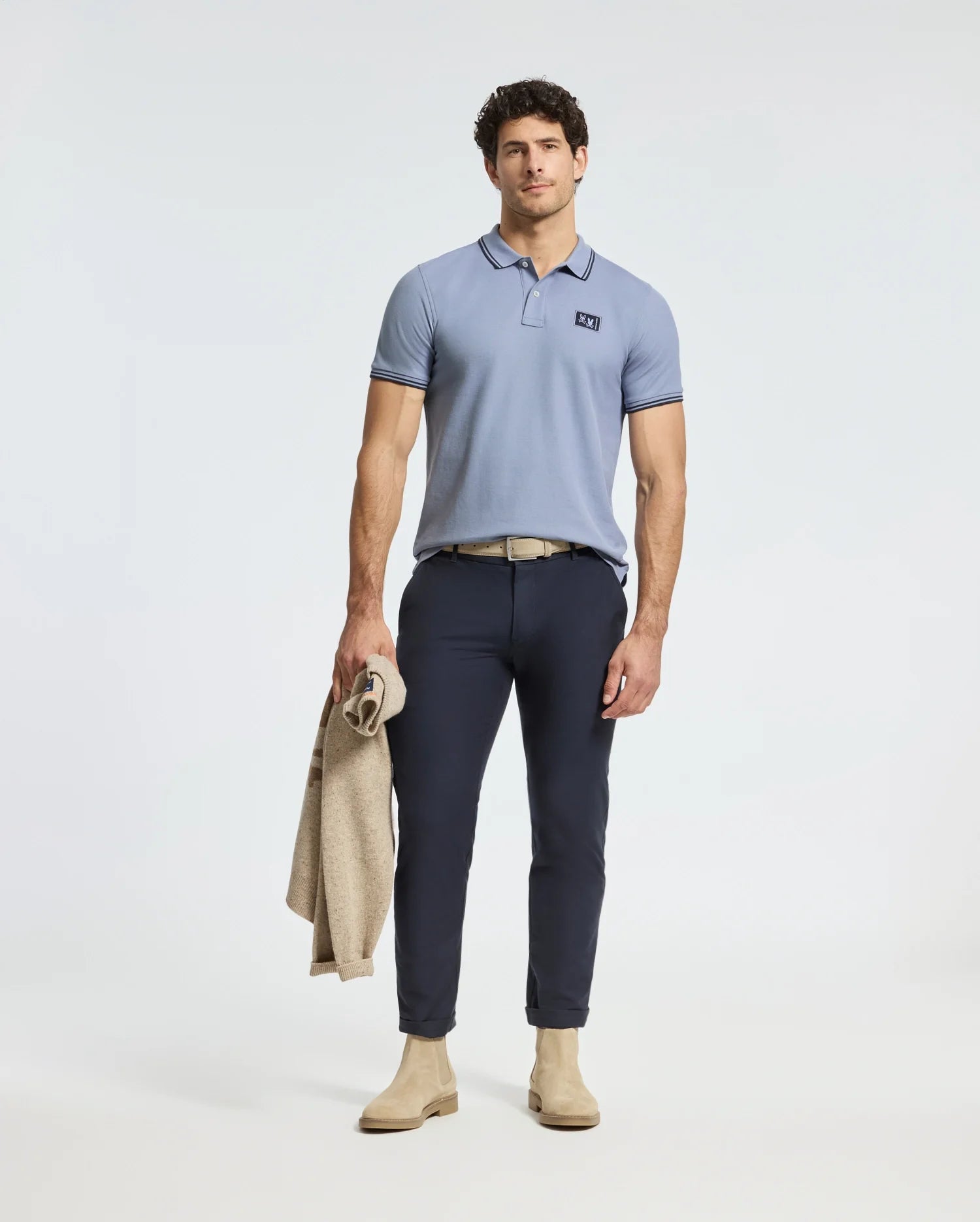A man in a Psycho Bunny MENS MONROE PIQUE POLO with Mother-of-Pearl buttons stands holding a beige jacket. He wears dark blue pants, a beige belt, and matching shoes, posing against a light background.