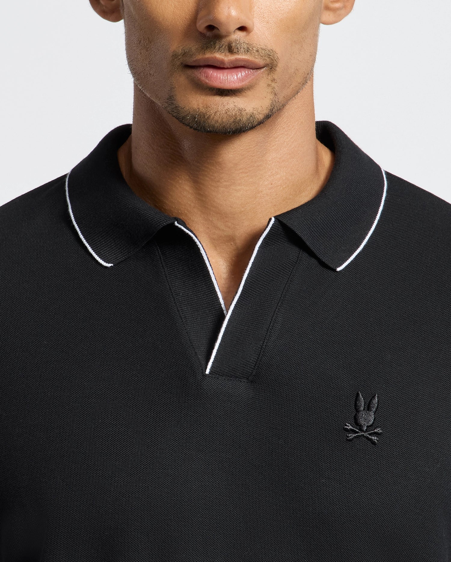 A person is wearing the MENS CREEDE PIQUE JOHNNY COLLAR POLO - B6M265E200 by Psycho Bunny. It is a men's long sleeve polo made from Pima cotton, featuring a black design with white piping and a distinctive Johnny collar. The left chest displays an embroidered logo of Psycho Bunny's signature stylized rabbit with crossed bones beneath, set against a plain white background.