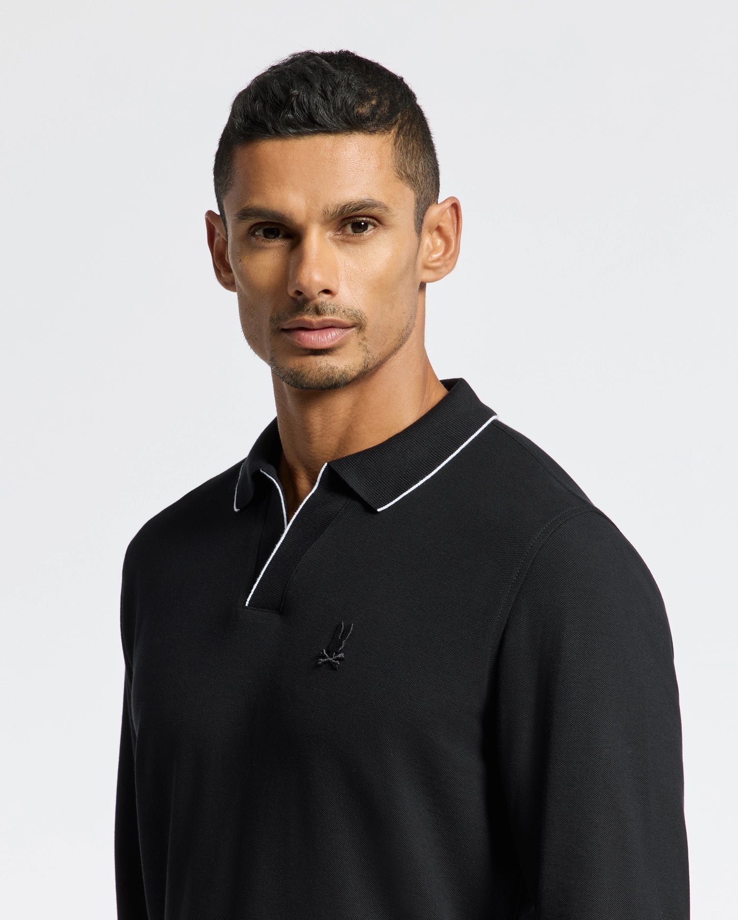 A person with short hair is wearing a black Psycho Bunny MENS CREEDE PIQUE JOHNNY COLLAR POLO - B6M265E200, which features a small embroidered logo on the chest. The Pima cotton shirt's collar has white piping, creating a subtle contrast against the plain, light-colored background.