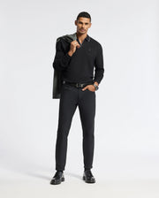 A man in a sleek black outfit poses confidently, showcasing a Psycho Bunny MENS CREEDE PIQUE JOHNNY COLLAR POLO alongside tailored pants and shoes. Adding a stylish touch, he casually drapes a jacket over his shoulder against a plain white background.