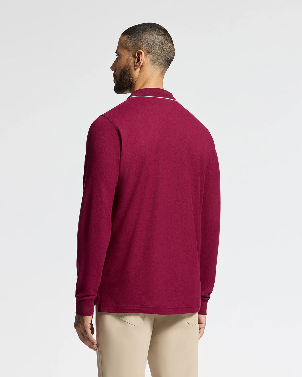 A person with short hair is facing away, wearing a maroon MENS CREEDE PIQUE JOHNNY COLLAR POLO by Psycho Bunny, crafted from soft Pima cotton, paired with beige pants. The background is plain and light-colored.
