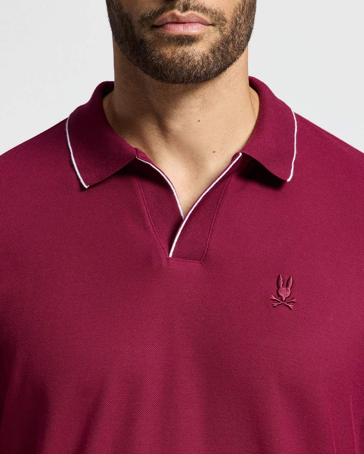 A man is wearing the Psycho Bunny MENS CREEDE PIQUE JOHNNY COLLAR POLO (B6M265E200) in burgundy, featuring a white-trimmed collar and an embroidered logo on the chest. The image highlights the refined details of this stylish shirt, focusing on the upper body.
