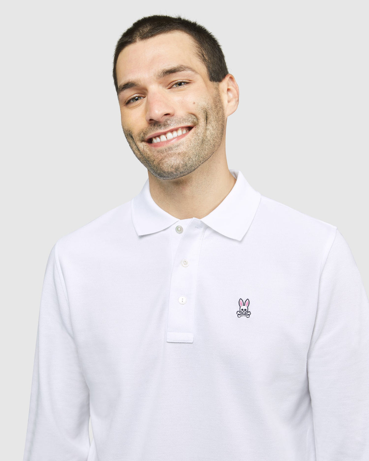 A smiling man with short dark hair and light stubble is wearing a Psycho Bunny MENS LONG SLEEVE CLASSIC POLO - B6M658ARPC made of 100% Pima cotton, featuring a small embroidered rabbit logo on the left side of the chest. The background is plain light gray.