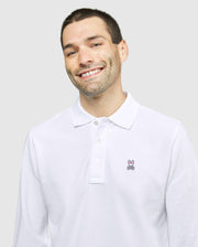 A smiling man with short dark hair and light stubble is wearing a Psycho Bunny MENS LONG SLEEVE CLASSIC POLO - B6M658ARPC made of 100% Pima cotton, featuring a small embroidered rabbit logo on the left side of the chest. The background is plain light gray.