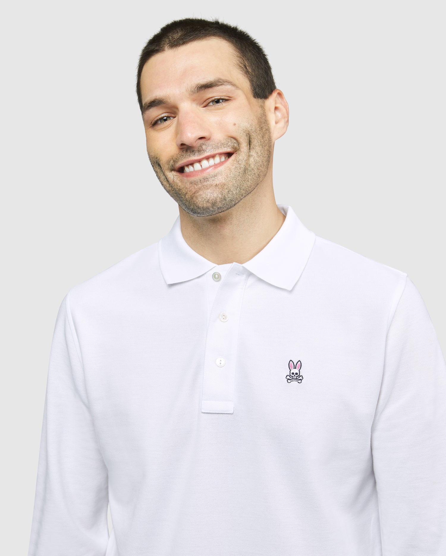A smiling man with short dark hair and light stubble is wearing a Psycho Bunny MENS LONG SLEEVE CLASSIC POLO - B6M658ARPC made of 100% Pima cotton, featuring a small embroidered rabbit logo on the left side of the chest. The background is plain light gray.