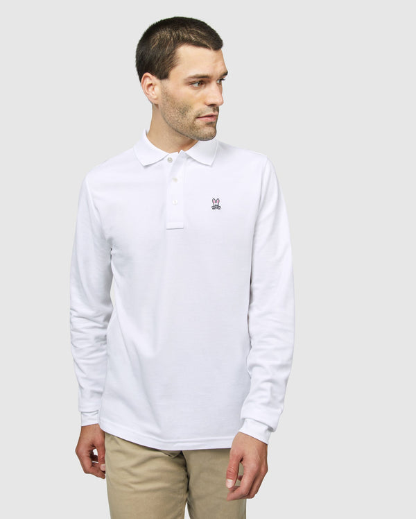 A man with short brown hair is wearing a long-sleeved white Men's Long Sleeve Classic Polo by Psycho Bunny (model B6M658ARPC), made from 100% Pima cotton, along with light beige pants. He is standing against a plain gray background, looking slightly to his left. There is a small embroidered logo on the left side of his shirt.