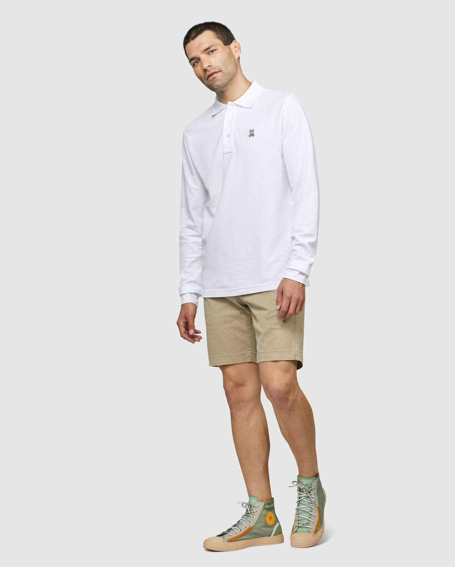 A man stands against a plain background wearing a Psycho Bunny MENS LONG SLEEVE CLASSIC POLO - B6M658ARPC in white, along with beige shorts and green high-top sneakers. He has short hair and is looking slightly downward with his hands relaxed by his sides.