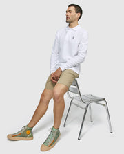 A person is seated on the edge of a silver metal chair, wearing a Psycho Bunny MENS LONG SLEEVE CLASSIC POLO - B6M658ARPC made of 100% Pima cotton in white, beige shorts, and green high-top sneakers. The background is plain and light-colored. The person gazes to the side with a relaxed posture.