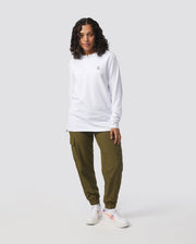 A person with curly hair is standing against a plain background. They are wearing the Psycho Bunny MENS LONG SLEEVE CLASSIC POLO - B6M658ARPC, a white shirt made from 100% Pima cotton featuring a small logo. They have on olive-green cargo pants and white sneakers with red accents. Their hands are at their sides, and they are looking at the camera.