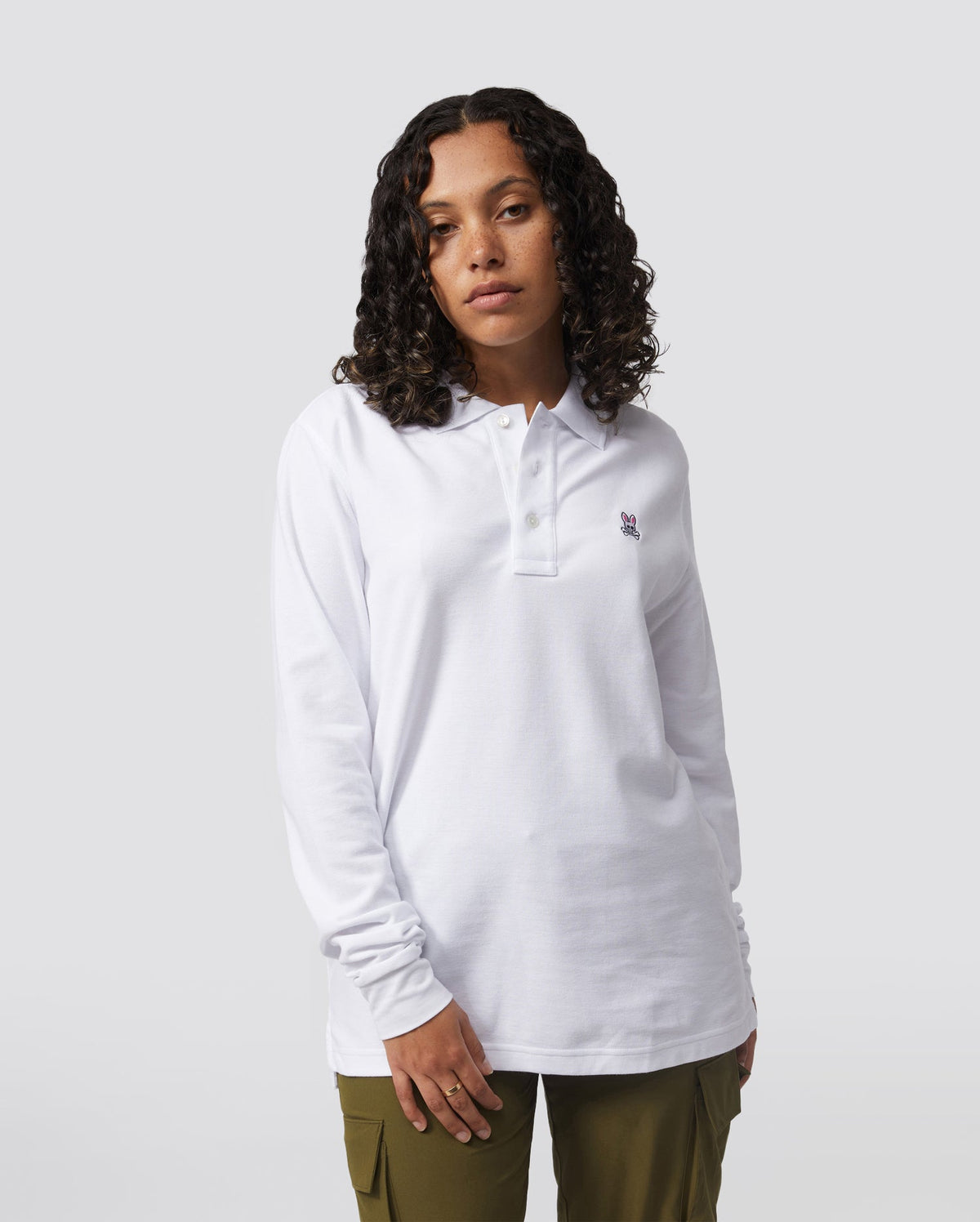 A person with curly hair is wearing a Psycho Bunny MENS LONG SLEEVE CLASSIC POLO (B6M658ARPC) made from 100% Pima cotton, paired with olive green pants. They are standing against a plain gray background, looking directly at the camera with a neutral expression.