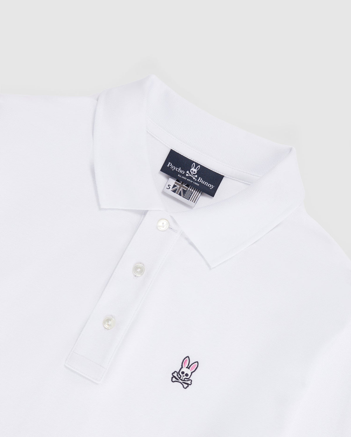 Introducing the MENS LONG SLEEVE CLASSIC POLO - B6M658ARPC from Psycho Bunny. This men's white polo shirt features a classic collar and three-button placket on the front, crafted from 100% Pima cotton in a diamond-knit piqué fabric for added texture. The left chest area displays a small, embroidered pink bunny with black crossbones logo. An inner neck label reads "Psycho Bunny." The backdrop is light gray.