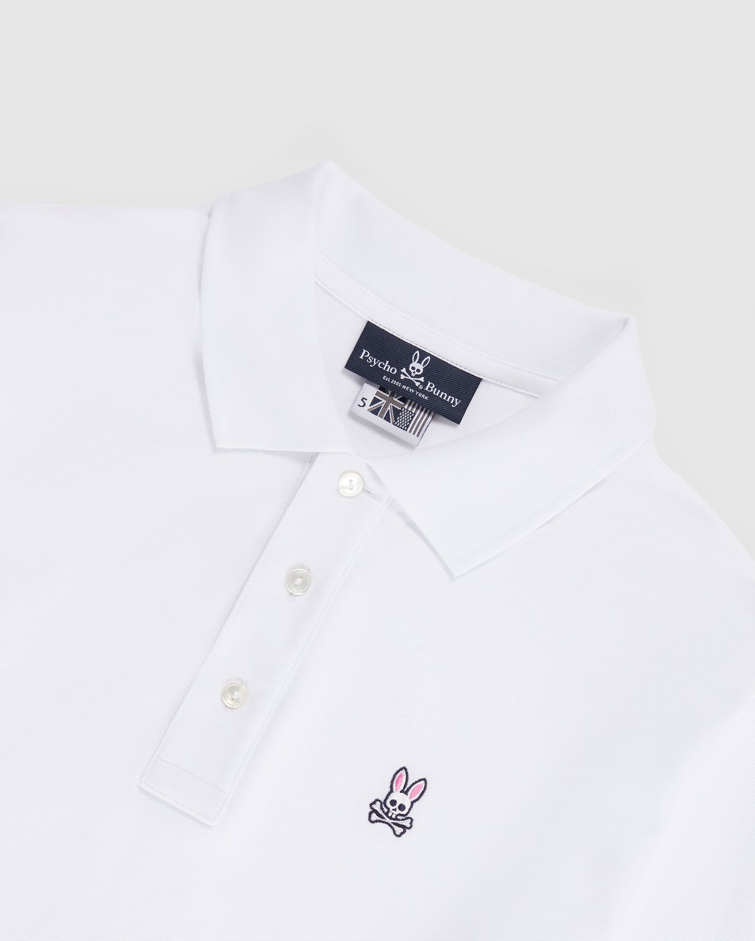 Introducing the MENS LONG SLEEVE CLASSIC POLO - B6M658ARPC from Psycho Bunny. This men's white polo shirt features a classic collar and three-button placket on the front, crafted from 100% Pima cotton in a diamond-knit piqué fabric for added texture. The left chest area displays a small, embroidered pink bunny with black crossbones logo. An inner neck label reads "Psycho Bunny." The backdrop is light gray.