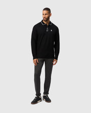 A man with short hair and a beard poses against a simple backdrop, wearing the Psycho Bunny MENS KENT LONG SLEEVE PIQUE POLO (B6M935D200), made from Pima cotton. The black shirt, accented with white trim and an emblem, effortlessly complements his dark pants and sneakers. His left hand is relaxed by his side while his right hand rests on his thigh.