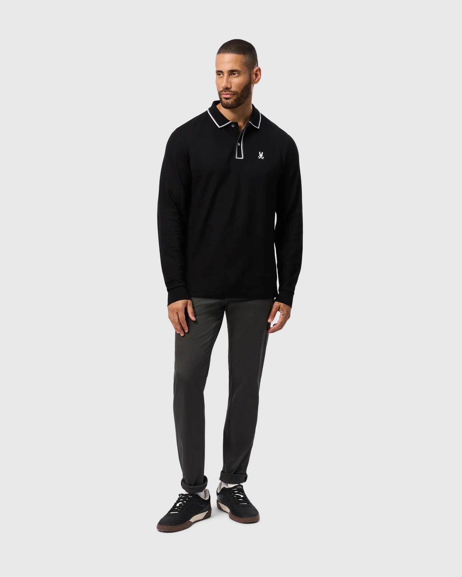 A man with short hair and a beard poses against a simple backdrop, wearing the Psycho Bunny MENS KENT LONG SLEEVE PIQUE POLO (B6M935D200), made from Pima cotton. The black shirt, accented with white trim and an emblem, effortlessly complements his dark pants and sneakers. His left hand is relaxed by his side while his right hand rests on his thigh.