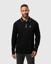 A man wearing the Psycho Bunny Kent Long Sleeve Pique Polo in black, featuring white trim and a small logo on the chest, is standing and smiling against a plain grey background. This polo, crafted from soft Pima cotton, demonstrates both style and comfort.