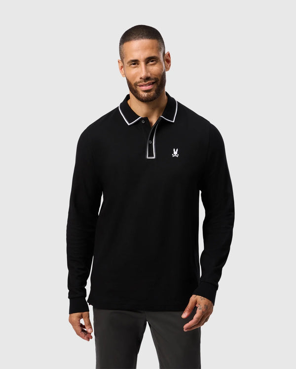 A man wearing the Psycho Bunny Kent Long Sleeve Pique Polo in black, featuring white trim and a small logo on the chest, is standing and smiling against a plain grey background. This polo, crafted from soft Pima cotton, demonstrates both style and comfort.
