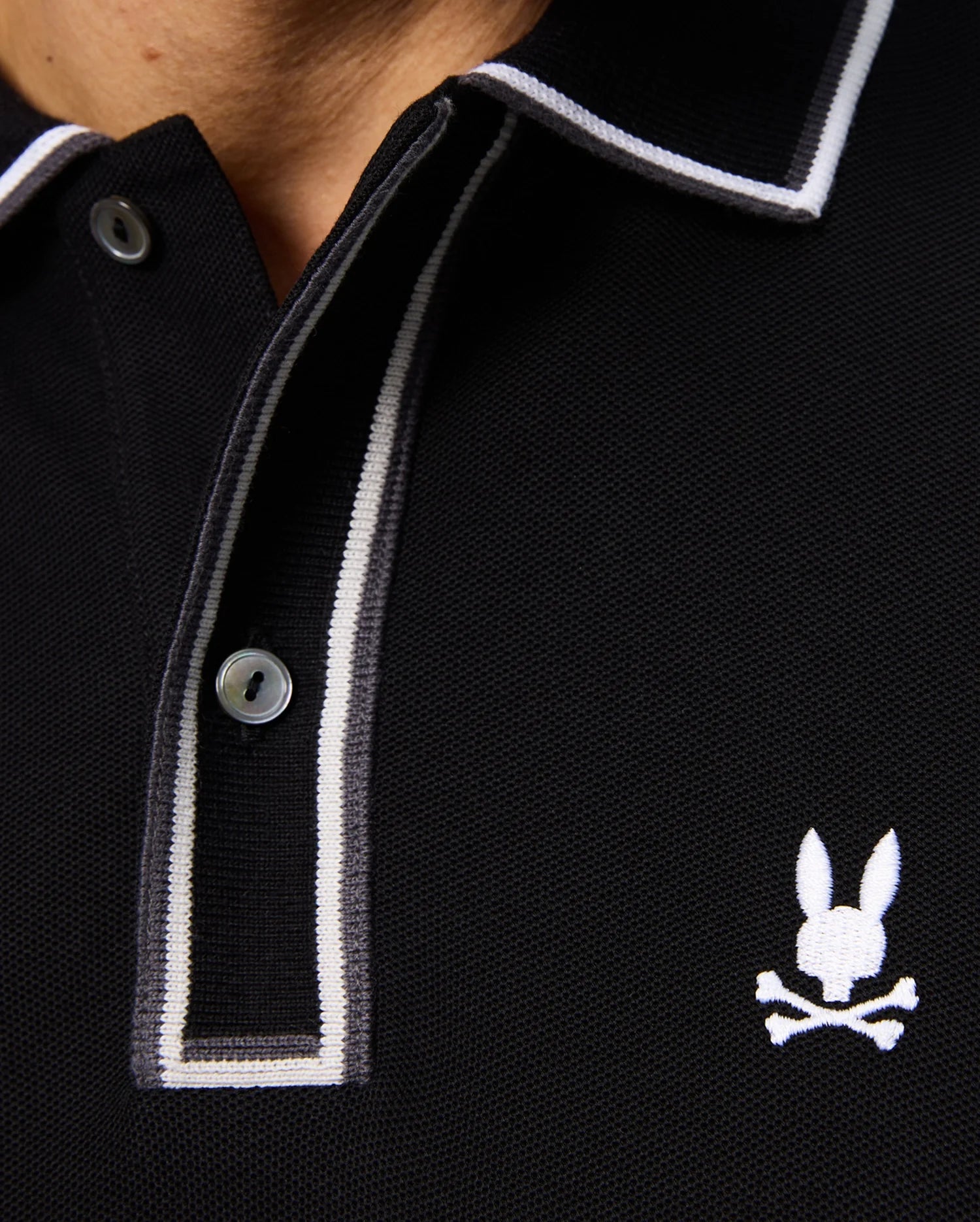 Close-up of the Psycho Bunny men's Kent long sleeve pique polo, style B6M935D200, in black with white and gray trim on the collar and button placket. Made from luxurious Pima cotton, it showcases an embroidered logo featuring a stylized white bunny head above crossed bones on the chest.