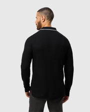 A man wearing the MENS KENT LONG SLEEVE PIQUE POLO - B6M935D200 by Psycho Bunny stands with his back to the camera against a light gray background. The rugby-style shirt features a collar with white trim. He has short hair and a beard, and his hands rest casually at his sides.