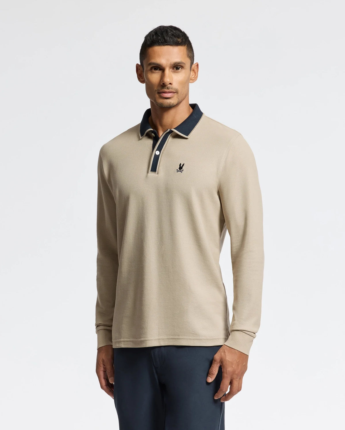 A man embodies understated elegance in the Psycho Bunny MENS KENT LONG SLEEVE PIQUE POLO (B6M935D200), a beige masterpiece made from pima cotton, accented with a dark collar and emblem. He poses against a simple backdrop, his short, dark hair flawlessly enhancing the outfit which is paired with dark pants.