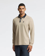 A man embodies understated elegance in the Psycho Bunny MENS KENT LONG SLEEVE PIQUE POLO (B6M935D200), a beige masterpiece made from pima cotton, accented with a dark collar and emblem. He poses against a simple backdrop, his short, dark hair flawlessly enhancing the outfit which is paired with dark pants.