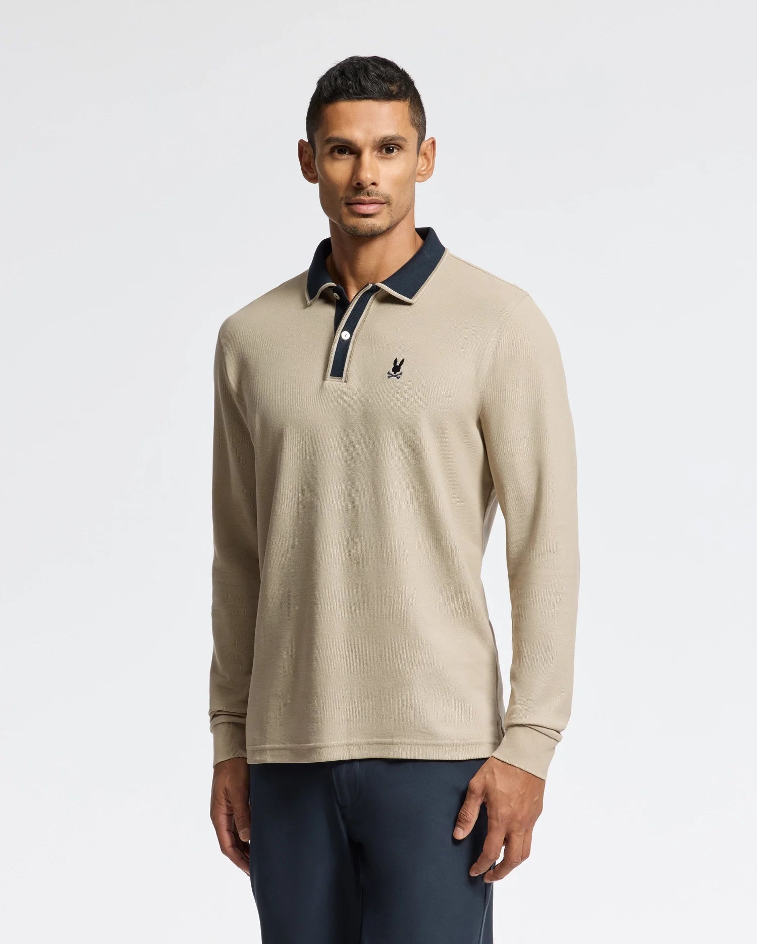 A man embodies understated elegance in the Psycho Bunny MENS KENT LONG SLEEVE PIQUE POLO (B6M935D200), a beige masterpiece made from pima cotton, accented with a dark collar and emblem. He poses against a simple backdrop, his short, dark hair flawlessly enhancing the outfit which is paired with dark pants.