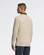 Dressed in a Psycho Bunny MENS KENT LONG SLEEVE PIQUE POLO in beige with a dark collar and paired with navy pants, this individual conveys understated luxury. They are standing and facing left, seen from the back against a plain white background.