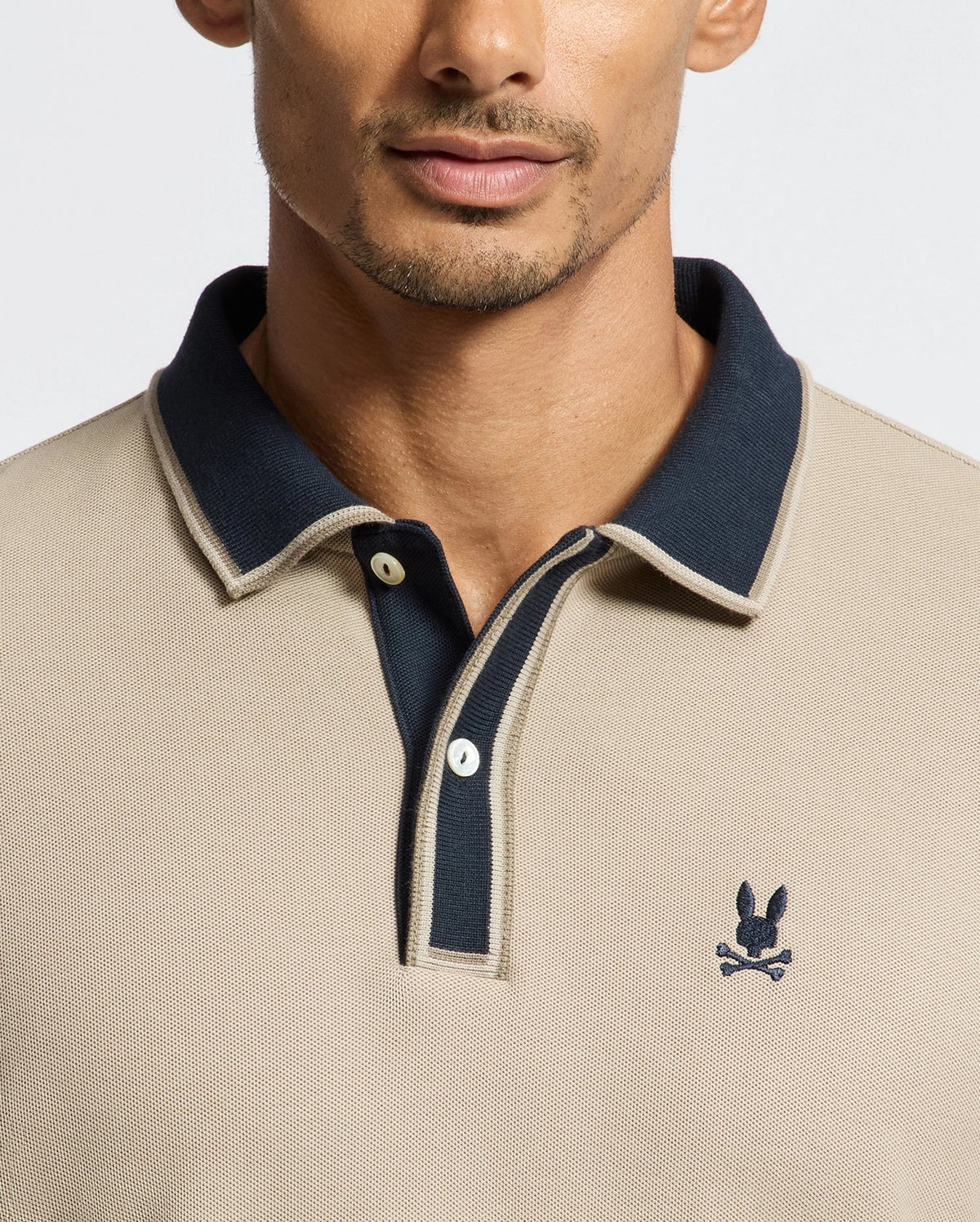 A man dressed in the MENS KENT LONG SLEEVE PIQUE POLO by Psycho Bunny, showcases quiet luxury with its beige Pima cotton fabric, accented by a dark collar and a subtle embroidered skull with rabbit ears logo on the chest.