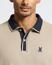 A man dressed in the MENS KENT LONG SLEEVE PIQUE POLO by Psycho Bunny, showcases quiet luxury with its beige Pima cotton fabric, accented by a dark collar and a subtle embroidered skull with rabbit ears logo on the chest.