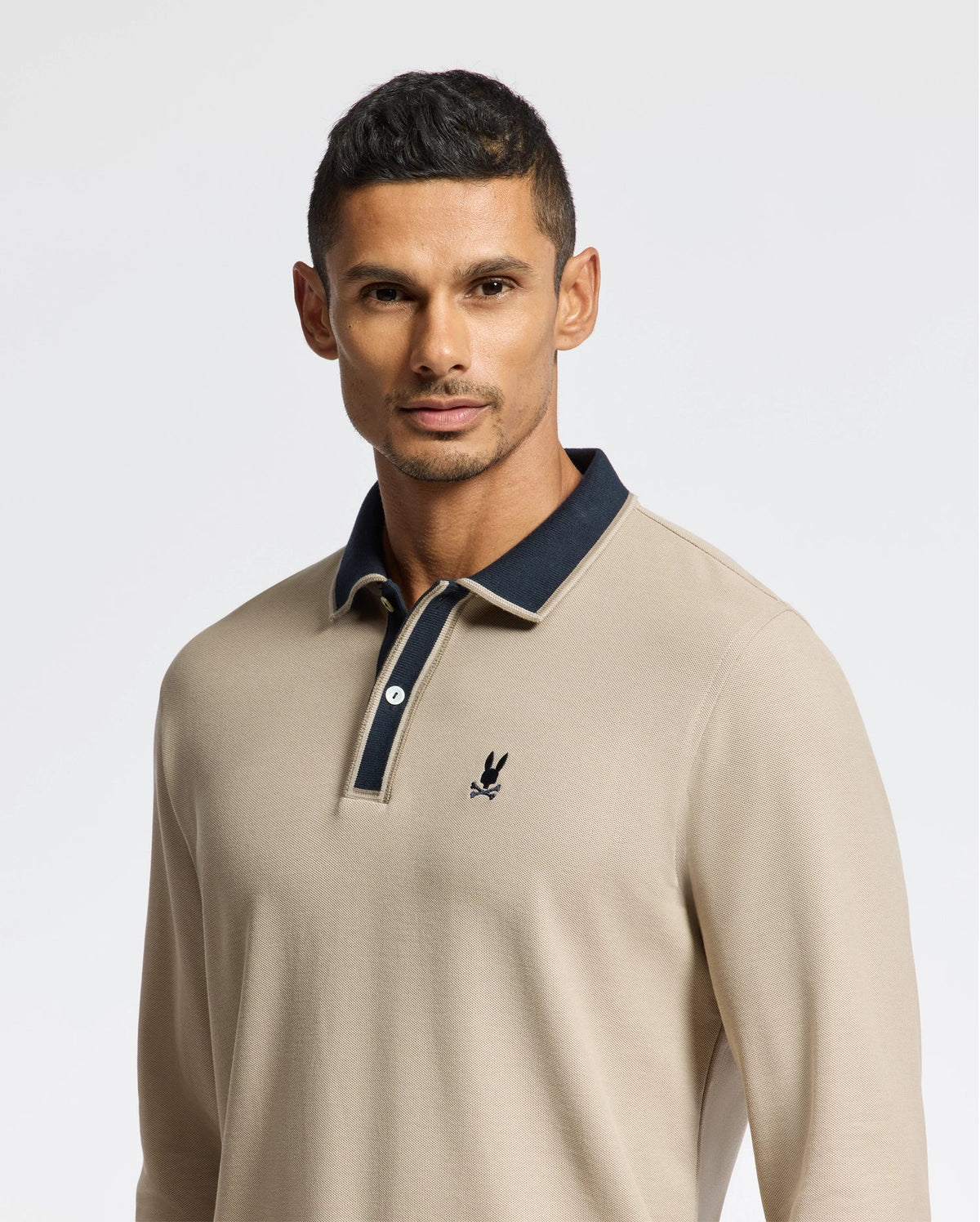 A man embodies understated elegance wearing the Psycho Bunny Men's Kent Long Sleeve Pique Polo (B6M935D200) in beige, made from Pima cotton. It showcases a dark collar with a discreet logo on the left chest. He stands against a light background, sporting short hair and a serene expression.