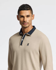 A man embodies understated elegance wearing the Psycho Bunny Men's Kent Long Sleeve Pique Polo (B6M935D200) in beige, made from Pima cotton. It showcases a dark collar with a discreet logo on the left chest. He stands against a light background, sporting short hair and a serene expression.