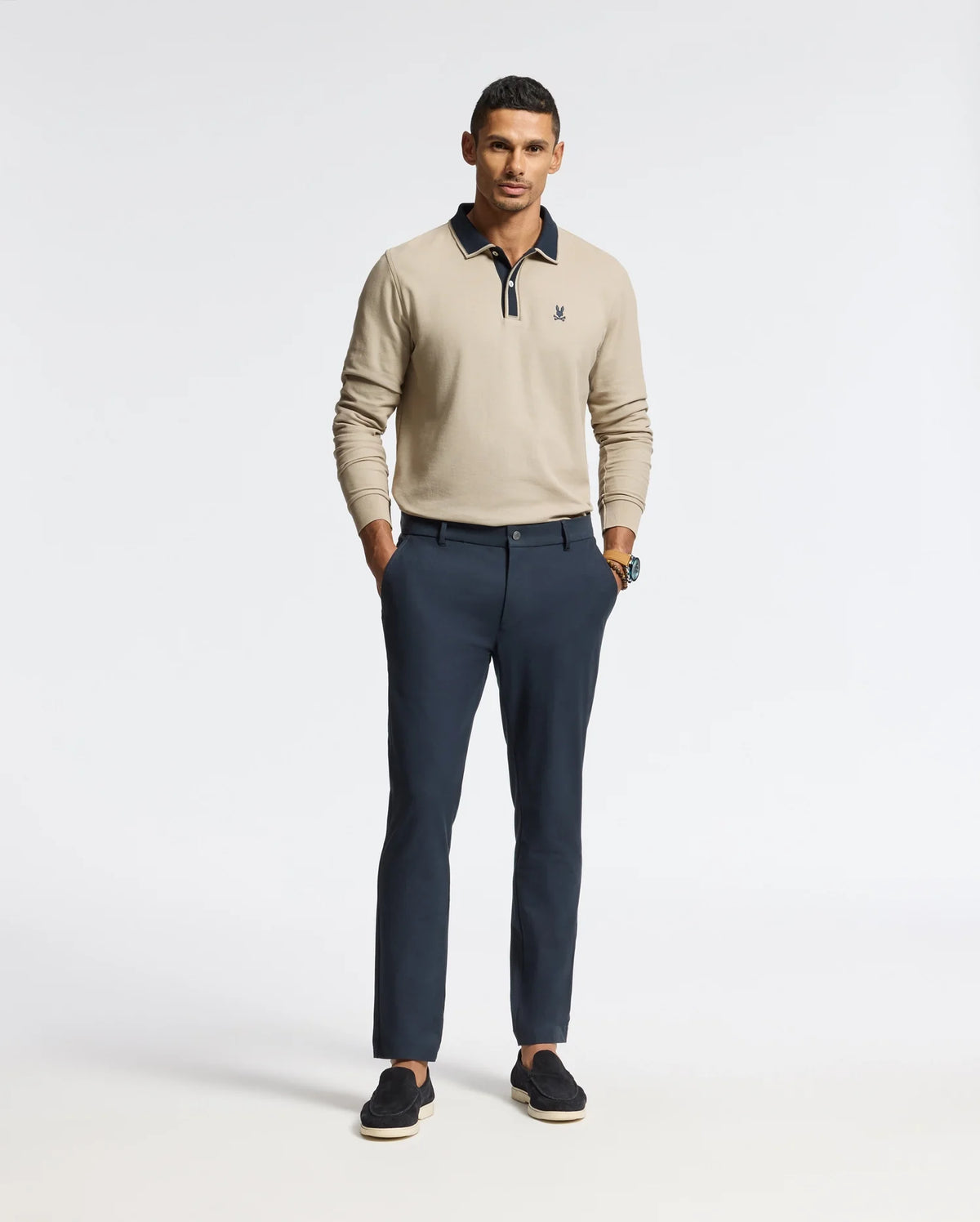 A man stands confidently in a Psycho Bunny MENS KENT LONG SLEEVE PIQUE POLO - B6M935D200 paired with dark blue trousers. With one hand in his pocket and wearing black loafers, he effortlessly exudes quiet luxury against the plain white background.