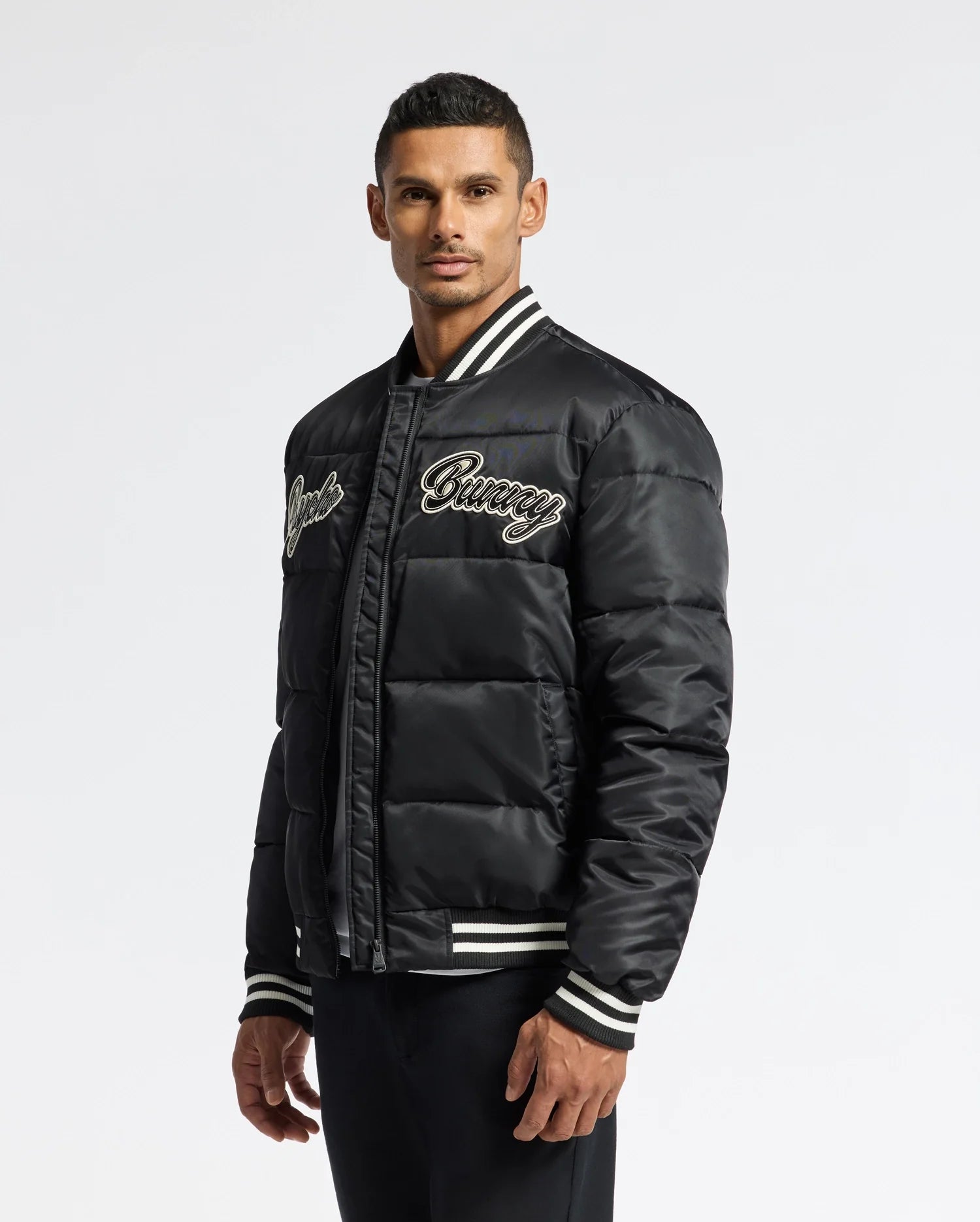 A man wearing the Psycho Bunny MENS FRAZER FASHION PUFFER, featuring white striped cuffs and embroidered text on the chest, stands against a plain background, looking at the camera.