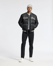 A man wearing a Psycho Bunny MENS FRAZER FASHION PUFFER - B6N338E200 stands against a plain white background. Hands in his pockets, he looks confidently at the camera, showcasing his stylish ensemble comprising a gray beanie, black varsity jacket, and white shirt with ease.