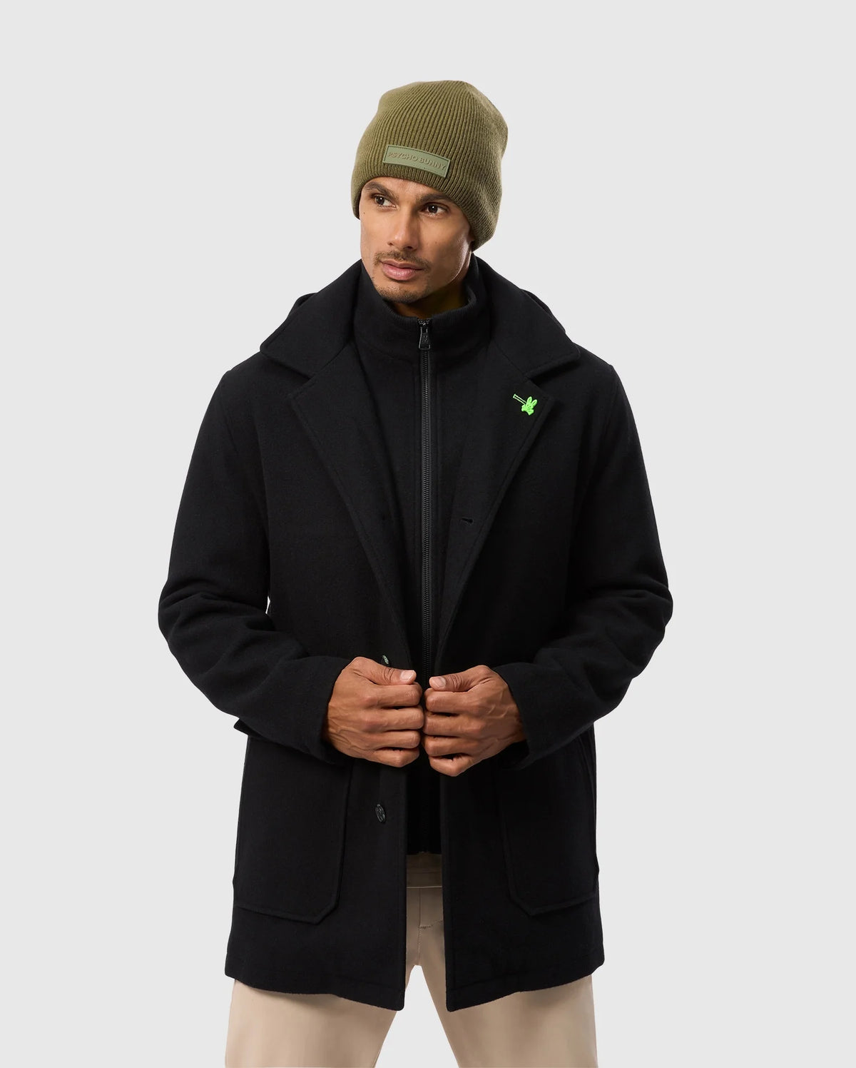 A person adjusts the front of their elegant Psycho Bunny MENS STAFFORD WOOL BLEND COAT - B6N551D200 against a light gray background, paired with a green beanie and beige pants.