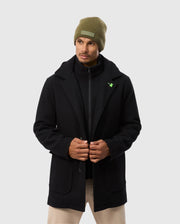 A person adjusts the front of their elegant Psycho Bunny MENS STAFFORD WOOL BLEND COAT - B6N551D200 against a light gray background, paired with a green beanie and beige pants.