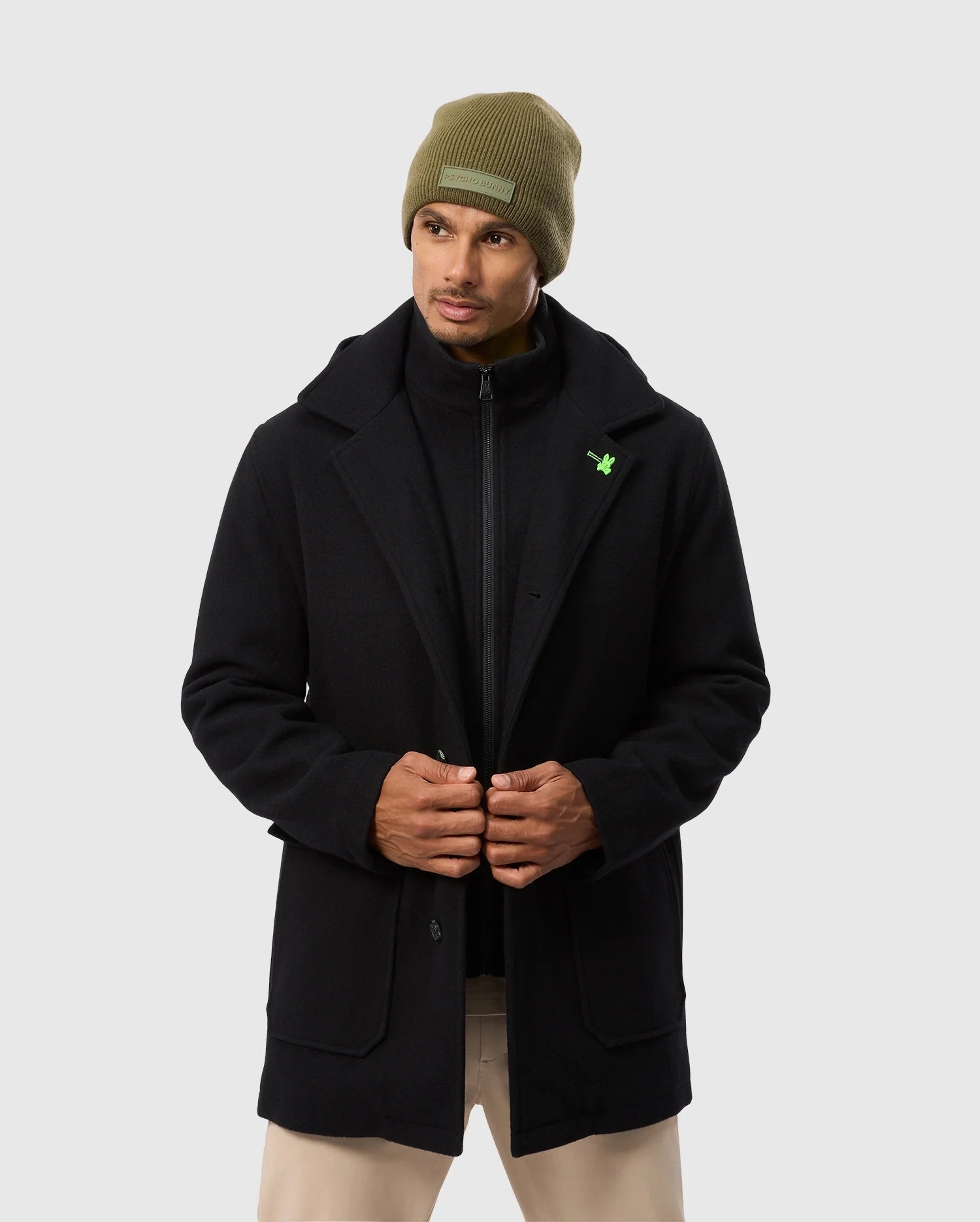 A person adjusts the front of their elegant Psycho Bunny MENS STAFFORD WOOL BLEND COAT - B6N551D200 against a light gray background, paired with a green beanie and beige pants.