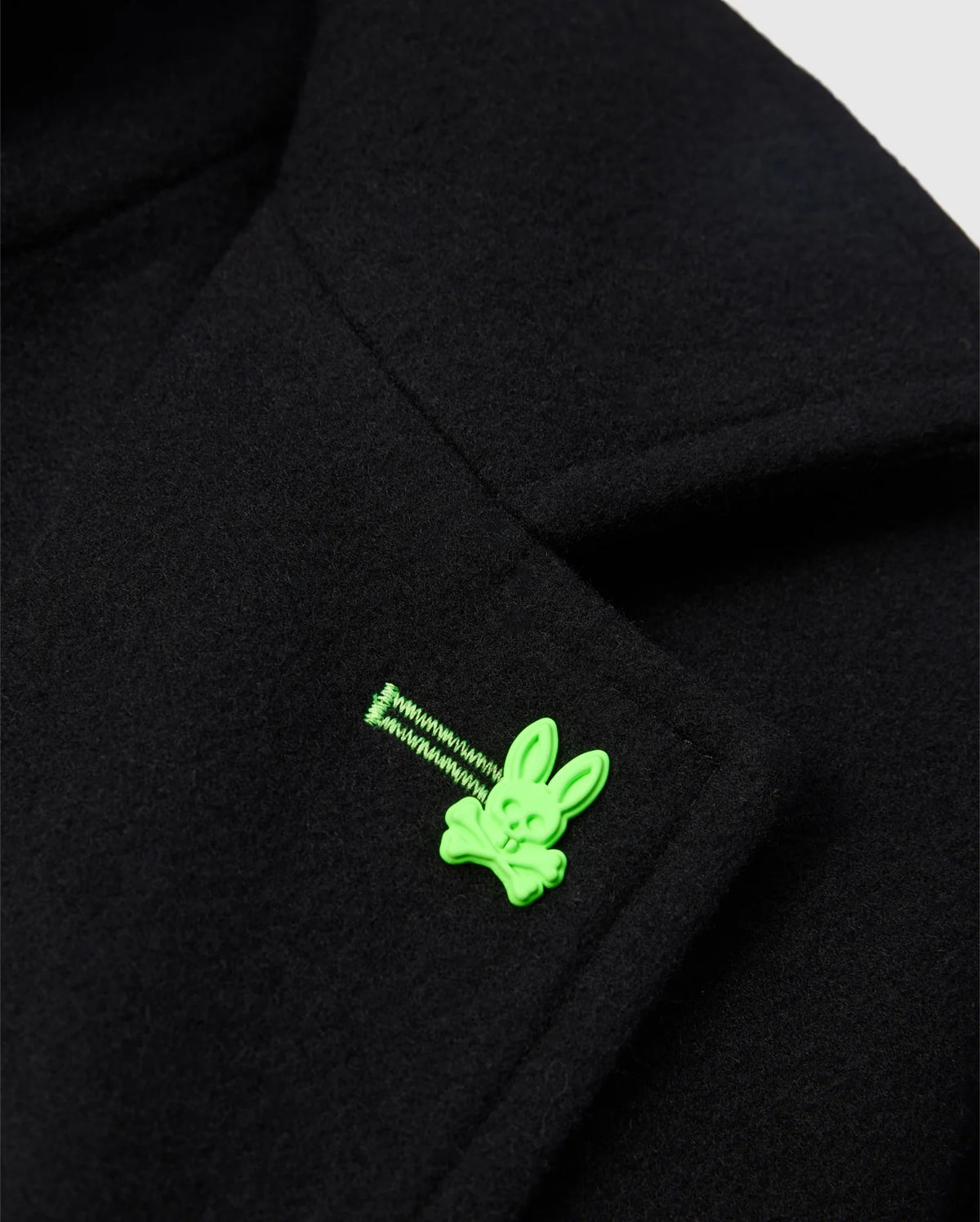 The MENS STAFFORD WOOL BLEND COAT - B6N551D200 from Psycho Bunny is an elegant black coat featuring a bright green enamel pin shaped like a bunny face above crossbones.