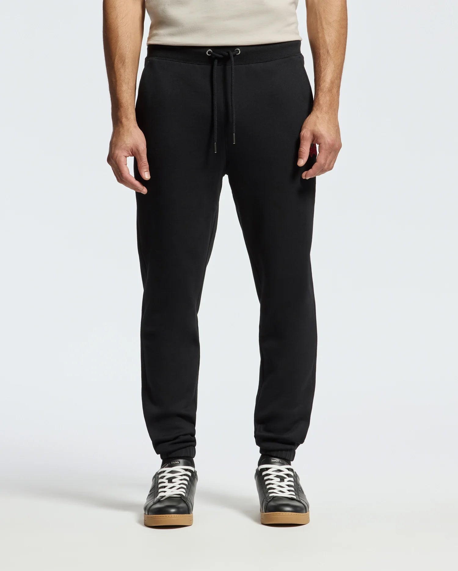 A person is wearing the Psycho Bunny MENS PIERCE ESSENTIAL FRENCH TERRY SWEATPANT in sleek black, complete with a drawstring waist, paired with black sneakers featuring white laces and soles. The background is plain white, and the upper body is partially visible.
