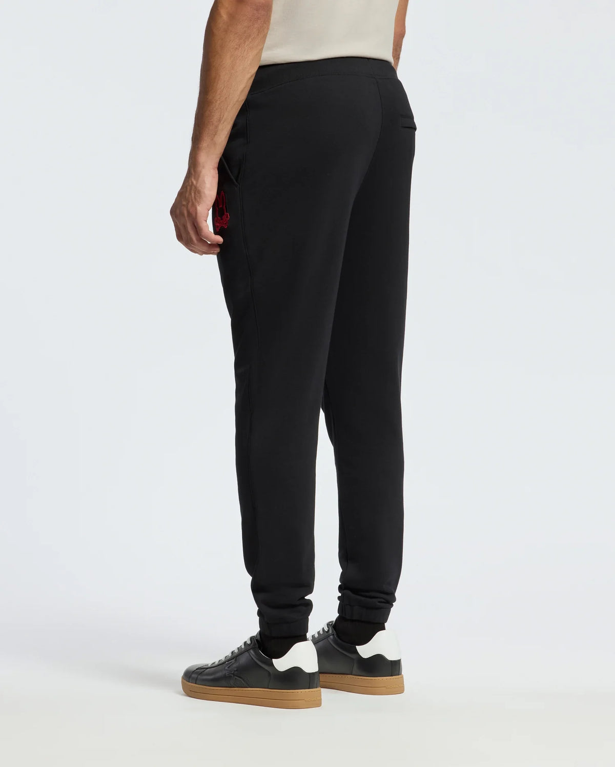 A person wearing the MENS PIERCE ESSENTIAL FRENCH TERRY SWEATPANT by Psycho Bunny stands facing away, paired with sleek black sneakers featuring white soles. The stylish black joggers, made from cozy French terry material, display a small red logo on the side against a plain white background.
