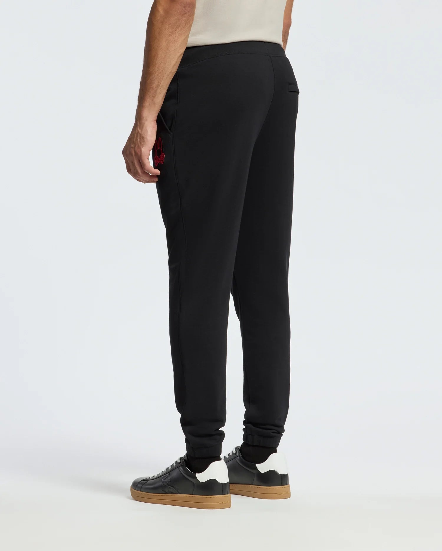 A person wearing the MENS PIERCE ESSENTIAL FRENCH TERRY SWEATPANT by Psycho Bunny stands facing away, paired with sleek black sneakers featuring white soles. The stylish black joggers, made from cozy French terry material, display a small red logo on the side against a plain white background.