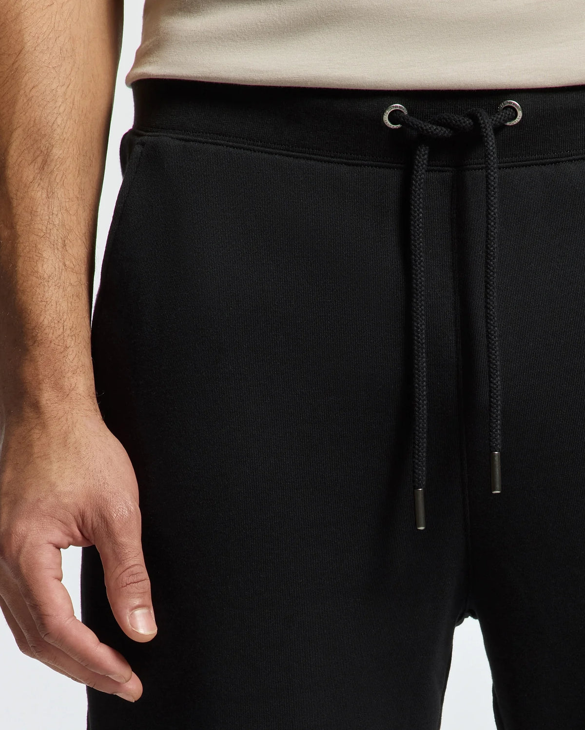 The image shows an individual dressed in Psycho Bunny's MENS PIERCE ESSENTIAL FRENCH TERRY SWEATPANT - B6P136E200 in black, paired with a beige top. The focus is on the waist area and part of the left arm, emphasizing the pants' lounge vibe with their front-hanging black drawstring featuring metal tips.