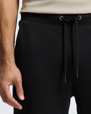 The image shows an individual dressed in Psycho Bunny's MENS PIERCE ESSENTIAL FRENCH TERRY SWEATPANT - B6P136E200 in black, paired with a beige top. The focus is on the waist area and part of the left arm, emphasizing the pants' lounge vibe with their front-hanging black drawstring featuring metal tips.