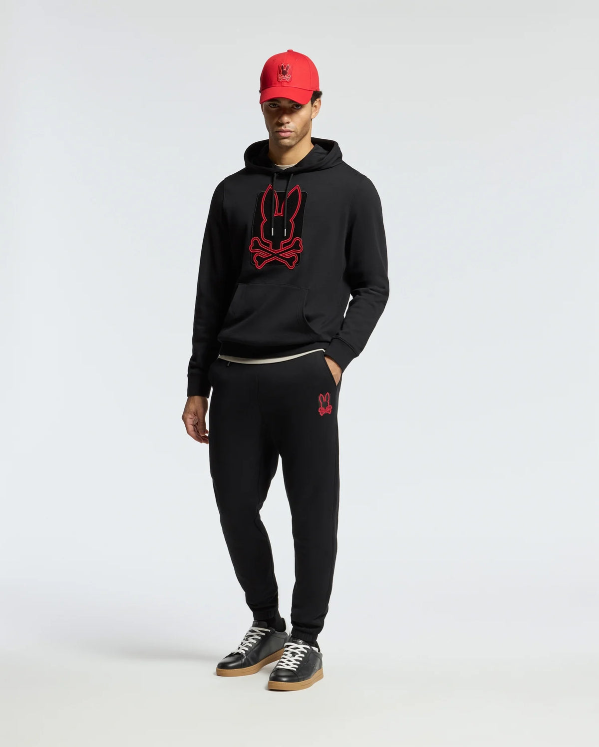 A person wearing a black hoodie with a red bunny logo, Psycho Bunny's MENS PIERCE ESSENTIAL FRENCH TERRY SWEATPANT - B6P136E200 featuring a small bunny design, and a red cap. They are standing against a plain white background and wearing black sneakers.