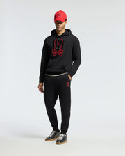 A person wearing a black hoodie with a red bunny logo, Psycho Bunny's MENS PIERCE ESSENTIAL FRENCH TERRY SWEATPANT - B6P136E200 featuring a small bunny design, and a red cap. They are standing against a plain white background and wearing black sneakers.